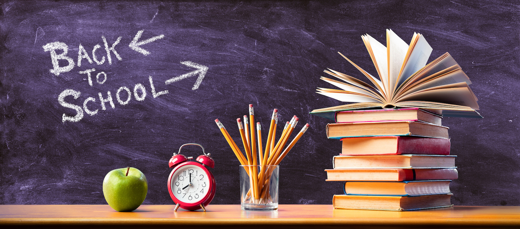 typical school items such as an apple pencils alarm clock and a stack of books are sitting on a wooden table in front of a chalkboard with back to school written on it in white chalk