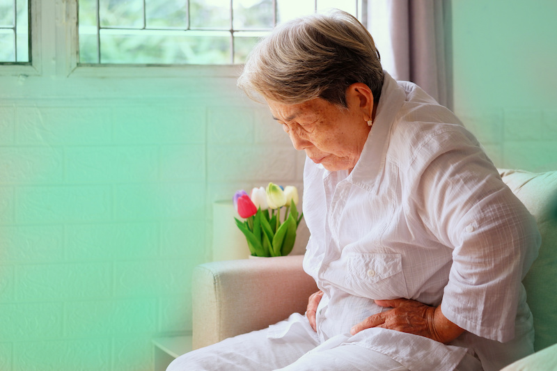 Elderly Asian woman with grey hair sits and holding her stomach in pain on the sofa