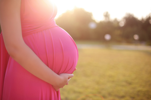 pregnant woman in pink shirt standing outside in a field resting her hands on her belly